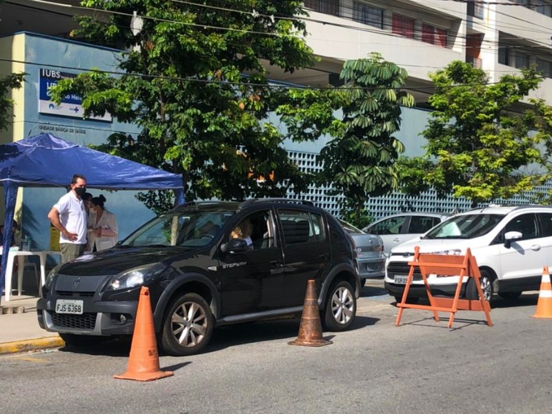 Um veículo preto estaciona no drive-thru montado na UBS Jardim Edite, numa tenda azul. Atrás dele tem um carro branco posicionado na fila